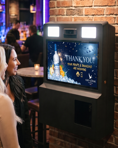 Illustrative bar scene with guests beside a wall-mounted photo booth displaying a thank-you message and sample Grey Goose branding.